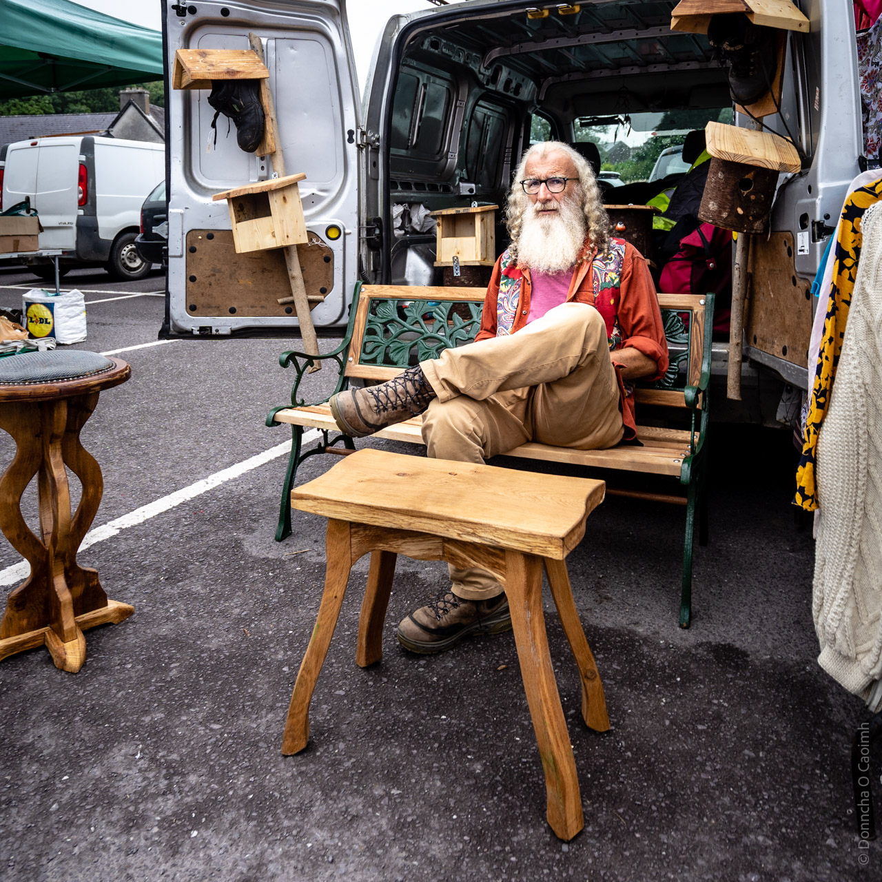 A craftsman sitting on a green metal and wooden bench beside his van at a market in Skibbereen, County Cork, surrounded by handmade wooden furniture and birdhouses, including a stool and table displayed in the foreground.