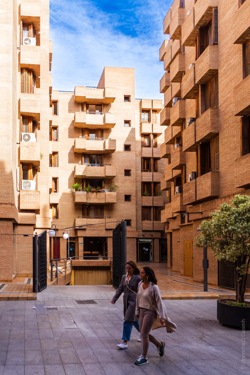 A modern residential building complex in Valencia, Spain, featuring brick architecture with protruding balconies, wooden window shutters, and air conditioning units. Two women are walking through the open courtyard, which has a paved pathway, a gated entrance, and potted trees. 