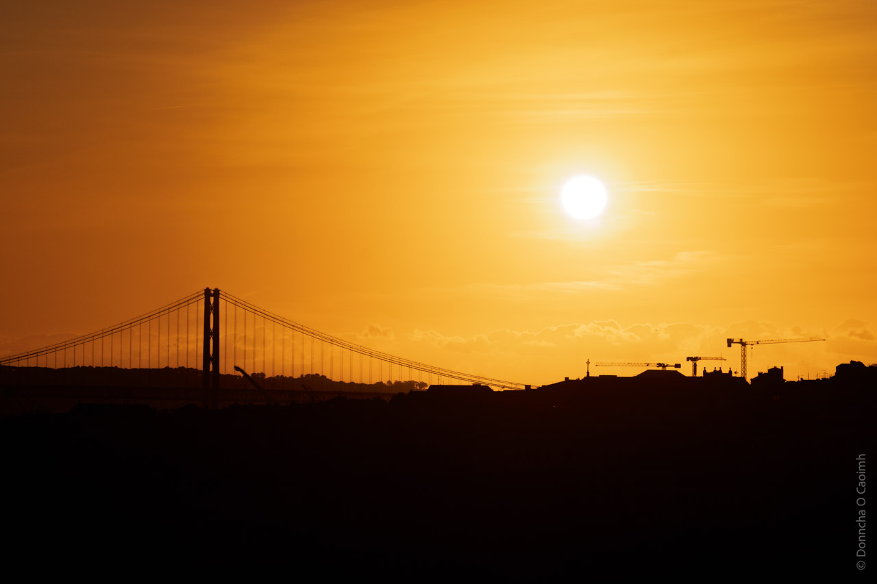 Silhouette of the 25 de Abril Bridge in Lisbon, Portugal, against a vibrant orange sunset sky with the sun glowing brightly; cranes and city structures visible in the foreground.

