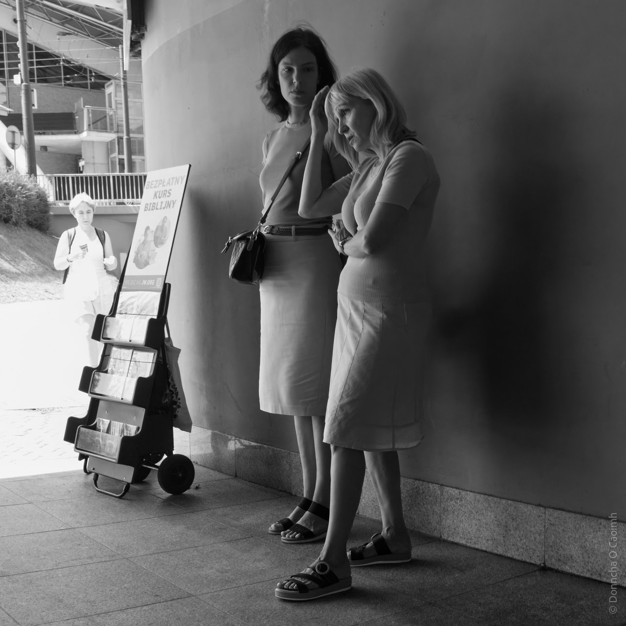 Black and white photo of two women standing near a wall in a metro station in Katowice, Poland. Both are dressed in light-coloured skirts and tops, wearing sandals, with one carrying a handbag. A literature stand displaying "Bezplatny Kurs Biblijny" (Free Bible Course) is visible nearby. Another person is walking in the background.