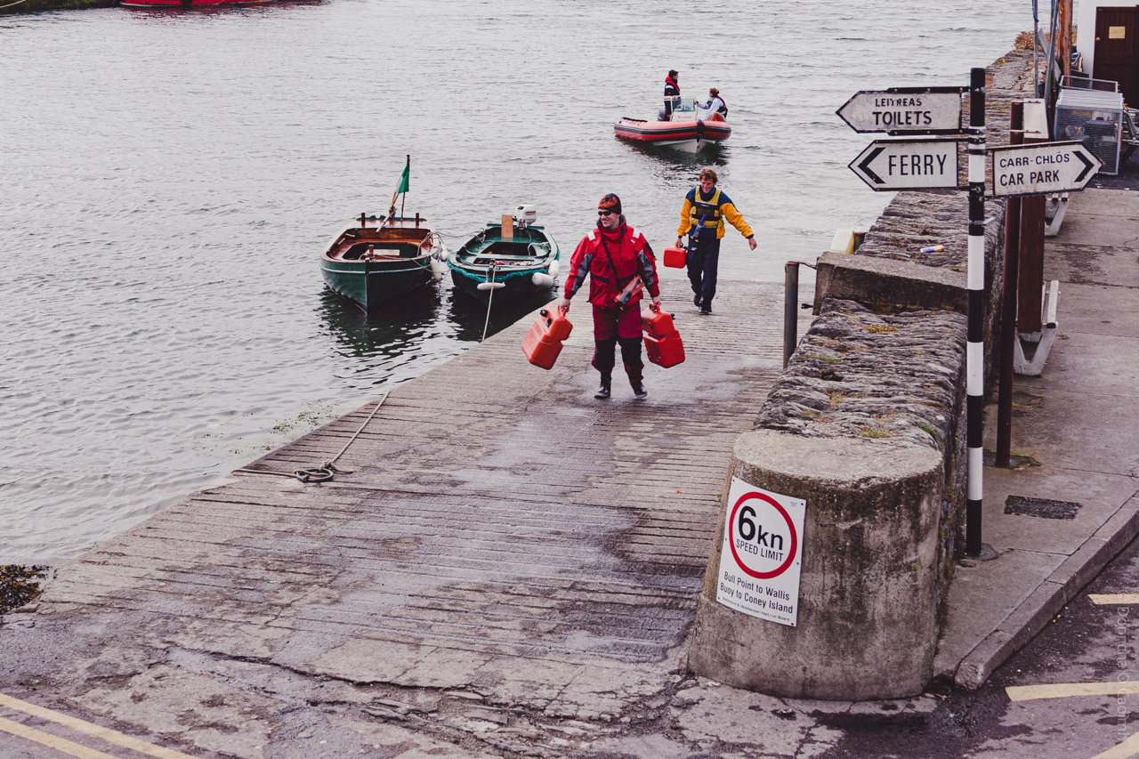 A harbor scene in Baltimore, Co Cork, Ireland showing people in waterproof gear carrying red fuel containers on a dock with small boats moored nearby, directional signs for "TOILETS", "FERRY", and "CAR PARK" visible, and a 6km speed limit sign in the foreground.