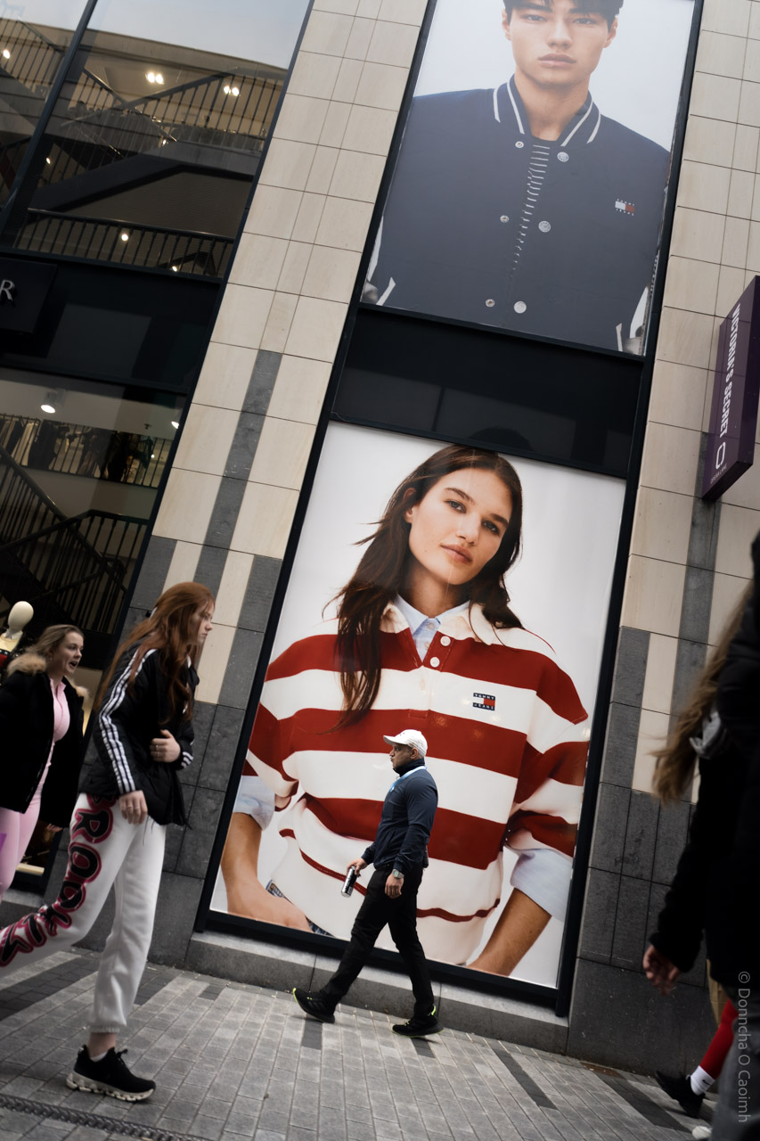 A candid street photograph taken in Cork City, Ireland, showing people walking past a large retail storefront with two oversized fashion posters. One poster features a person in a navy varsity jacket, and the other shows a person in a red and white striped rugby shirt, both displaying the Tommy Hilfiger logo. The scene captures urban life and the intersection of fashion advertising and everyday movement.