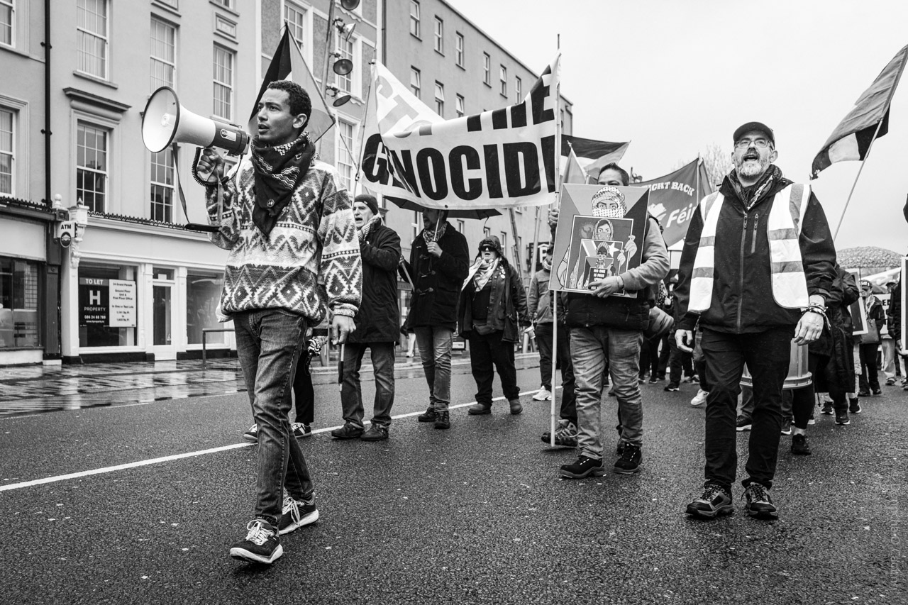 Black and white photo of a street protest in Cork, Ireland, supporting Gaza; people march holding banners and signs, including one reading "STOP THE GENOCIDE," with one person using a megaphone and another wearing a high-visibility vest, on a wet city street.
