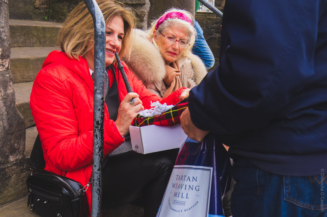A candid street scene in Edinburgh, Scotland, showing two women sitting on stone steps, one wearing a bright red jacket and holding a boxed junior bagpipe with a tartan cover, while a person in the foreground holds a "TARTAN WEAVING MILL" shopping bag.