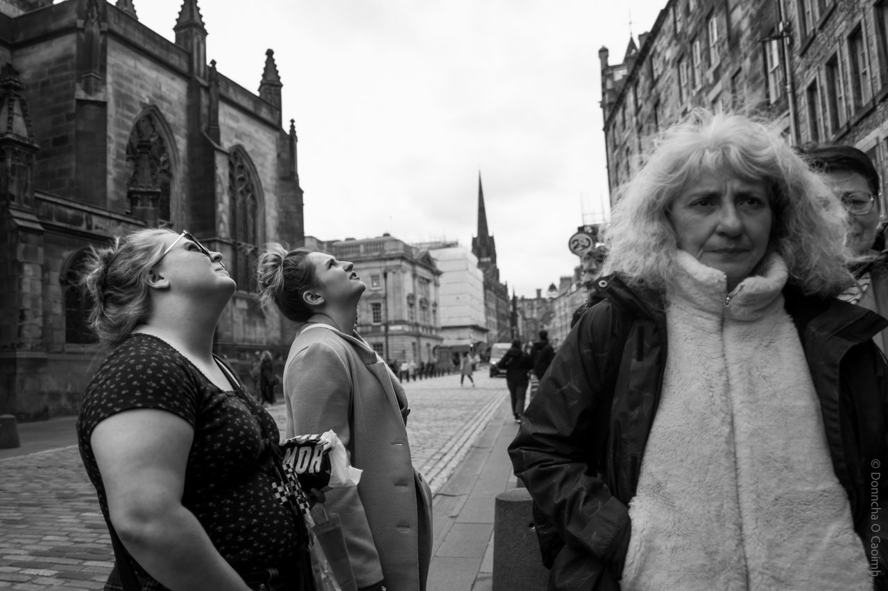 Black and white street photograph taken in Edinburgh, Scotland, showing several people standing and looking upwards on a cobblestone street, with historic stone buildings and a church featuring gothic architecture in the background; the image is candid and atmospheric, capturing a moment of curiosity or sightseeing.
