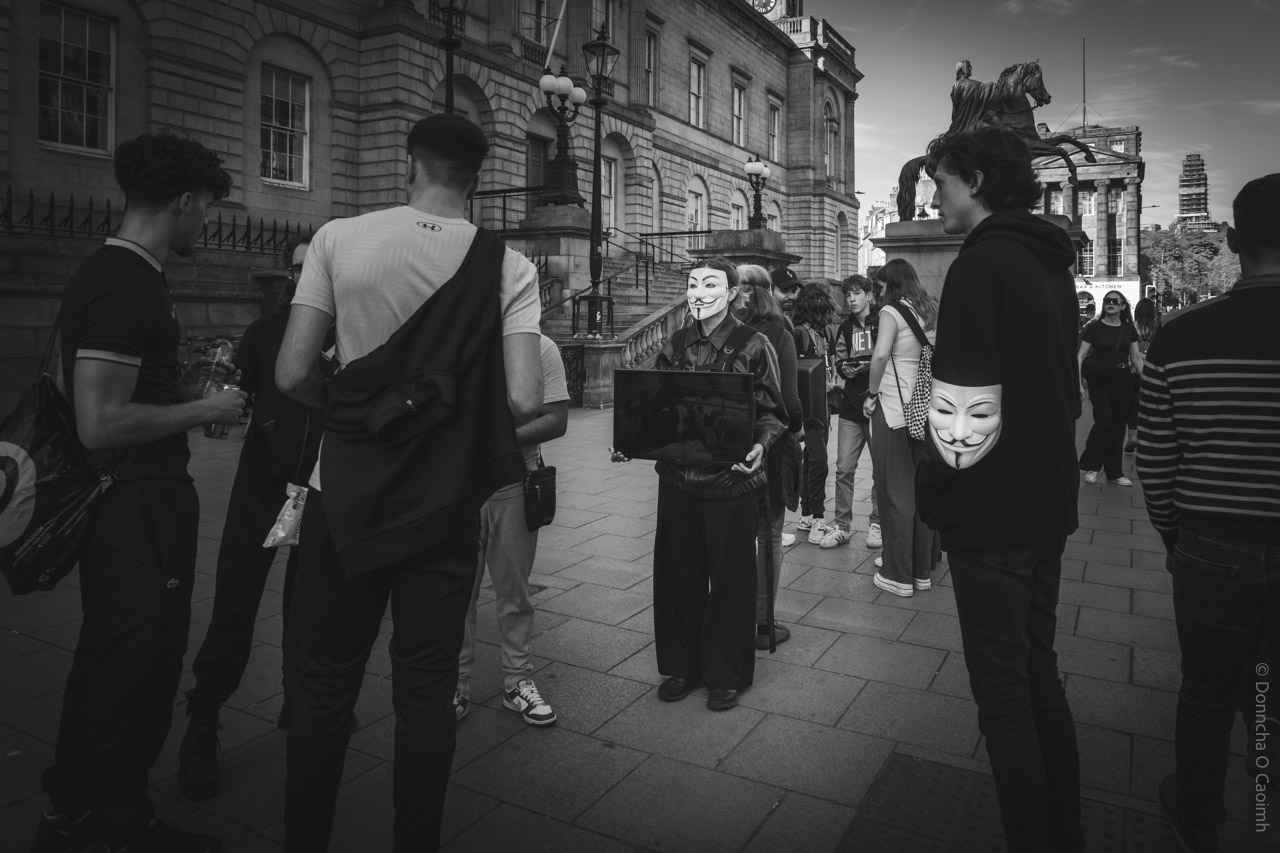 A black-and-white street photograph taken in Edinburgh, Scotland, showing a group of people gathered outside a historic building. Central in the image is a person wearing a Guy Fawkes mask, holding a screen as part of an "Anonymous for the Voiceless" demonstration. Several other people stand around, some engaged in conversation, with the statue of a horse and rider visible in the background.