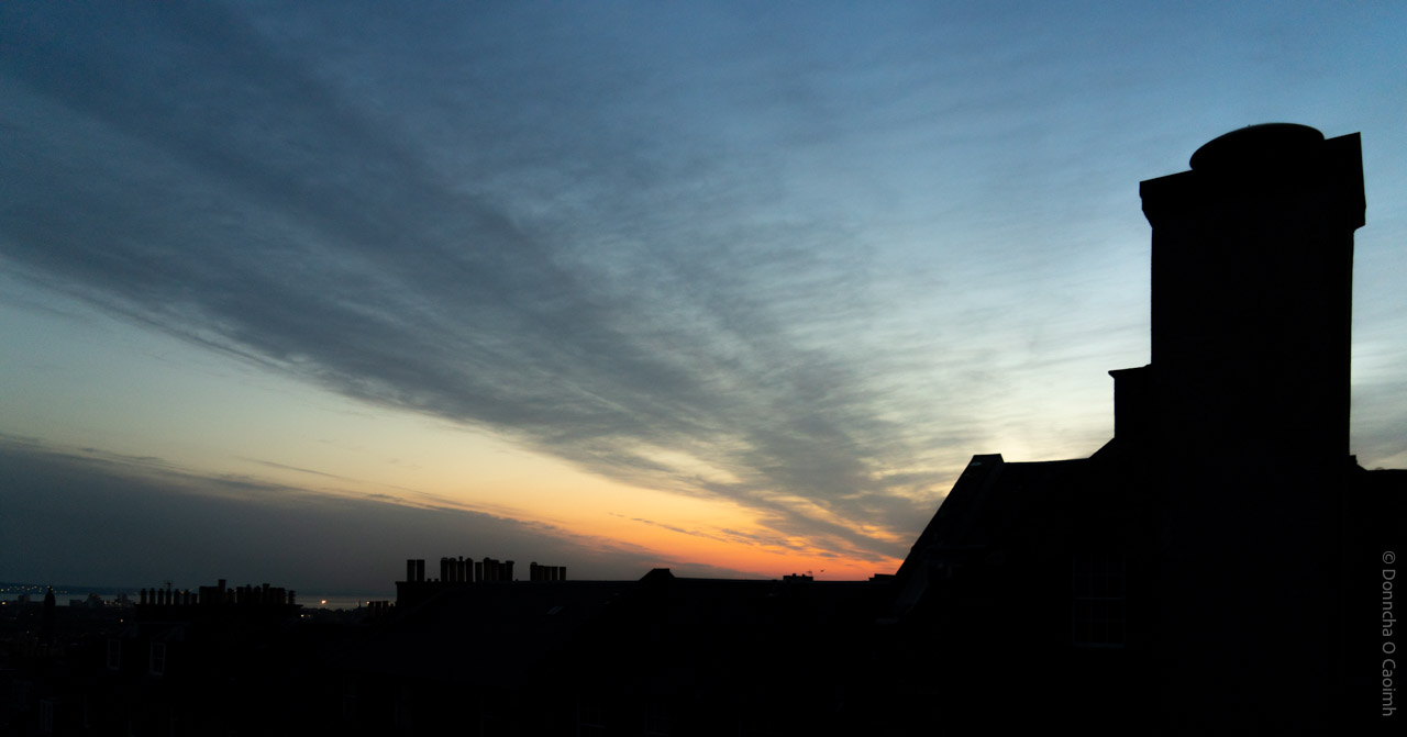 A silhouette of Edinburgh rooftops and chimneys at sunrise, with a dramatic sky streaked with clouds in blue, grey, and orange hues.