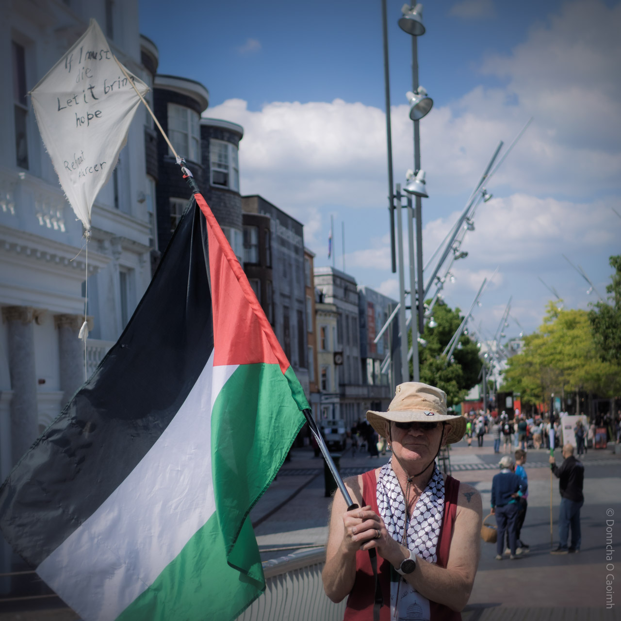 A person standing on a city street in Cork, Ireland, holding a large Palestinian flag and a kite. The kite has handwritten text: "If I must die Let it bring hope" and "
Refaat Alareer". The background shows a pedestrian area with people walking, modern streetlights, and historic buildings under a partly cloudy sky.
