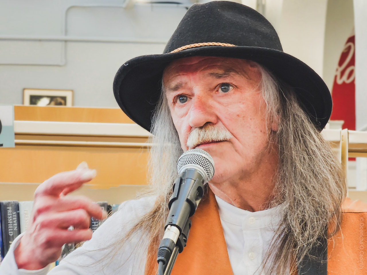 A detailed close-up of a musician in Cork City Library, wearing a black hat and brown vest, speaking into a microphone with bookshelves behind. The musician's long hair and expressive hand gesture are prominent.
