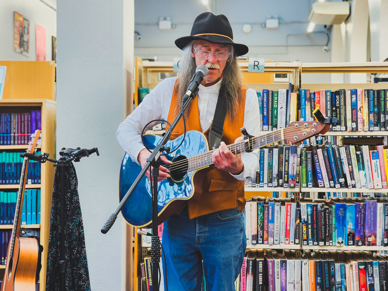 A close-up image of a musician playing a blue acoustic guitar and singing into a microphone in Cork City Library. The performer wears a black hat, white shirt, and brown vest, with bookshelves and a guitar on a stand in the background.

