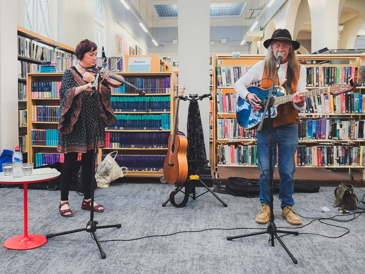 A candid photograph of two musicians performing in Cork City Library, surrounded by bookshelves. One musician plays the violin while the other plays an acoustic guitar and sings into a microphone. A small red table with water glasses and a bottle stands in the foreground, and a guitar rests on a stand between the performers. A library sign reading "Romance" is visible on a shelf behind them.
