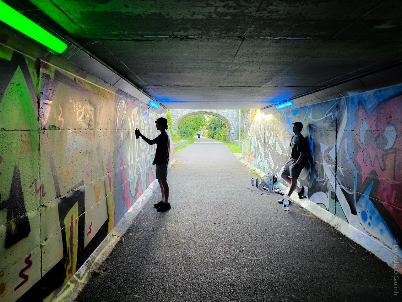 A vibrant urban scene in a Cork, Ireland underpass, featuring two people creating graffiti art on the tunnel walls under colourful neon lights, with spray paint cans scattered on the ground and lush greenery visible at the tunnel's exit.
