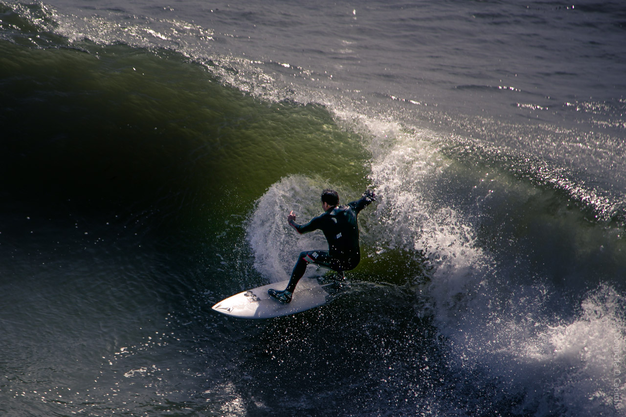 A surfer in a black wetsuit carves along the face of a large green wave in Santa Cruz, California, leaving a spray of water behind.

