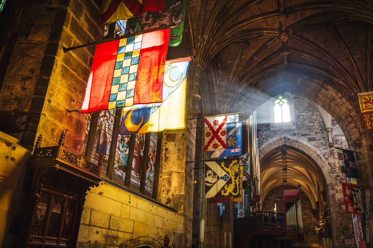 A vibrant interior view of St. Giles Cathedral in Edinburgh, showcasing medieval stone arches, colourful heraldic banners hanging from the walls, and sunlight streaming through stained glass windows, illuminating the historic architecture and ornate woodwork.
