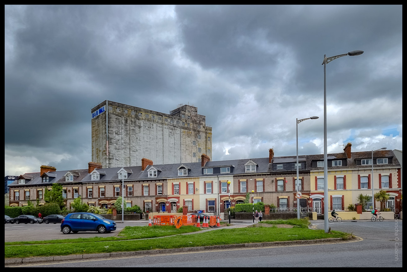 A colour photograph taken on Albert Road in Cork, Ireland, showing a row of Victorian terraced houses with red and cream facades in the foreground, a small car park and green verge, and a dramatic sky overhead. Behind the houses, the imposing, weathered concrete R&H Hall grain silo dominates the skyline, bearing the faded blue "R&H HALL" signage. The scene includes a few parked cars, some cyclists, pedestrians, and orange construction barriers. The R&H Hall building, a well-known industrial landmark, has since been demolished.