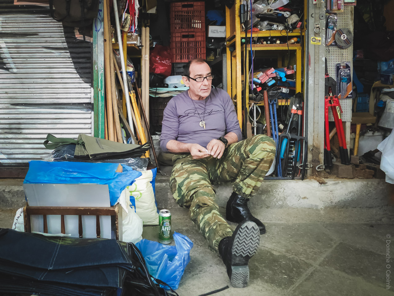 A candid street photo taken in Athens, Greece, showing a man in camouflage trousers and black boots sitting on the ground outside a cluttered tool shop, surrounded by bags, a can of Areti beer, and various hardware items, with shelves full of tools and equipment in the background.
