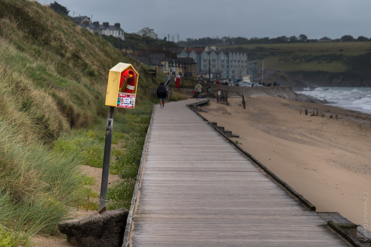 A wooden boardwalk runs alongside grassy sand dunes on a cloudy day in Youghal, Co Cork, Ireland. A yellow emergency lifebuoy station is mounted on a pole to the left of the boardwalk, with a red and white lifebuoy inside and a sign that reads "EMERGENCY LIFE BUOY" and "DIAL 999 OR 112." In the background, people walk along the boardwalk towards a row of pastel-coloured seaside houses, with the sandy beach and waves of the sea visible on the right.
