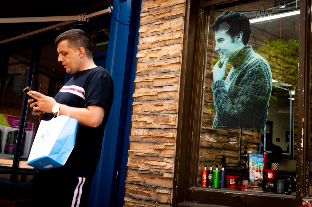 A candid street photo taken in Cork, Ireland, showing a person standing outside a shop with a blue and white shopping bag, looking at their phone. The shop has a brick façade and a large window displaying a poster of Elvis Presley in a chunky knit jumper. Various hair products, including colourful bottles and tubs, are lined up on the window ledge. The shop window features the text "CREW Official Supplier to Men".