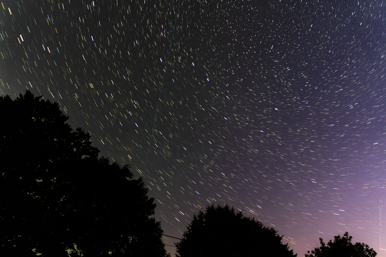 Long-exposure photograph of the night sky taken in Cork, Ireland, showing star trails forming circular arcs around the celestial pole, with silhouettes of trees and faint power lines at the bottom, and a subtle gradient of purple and black in the sky.