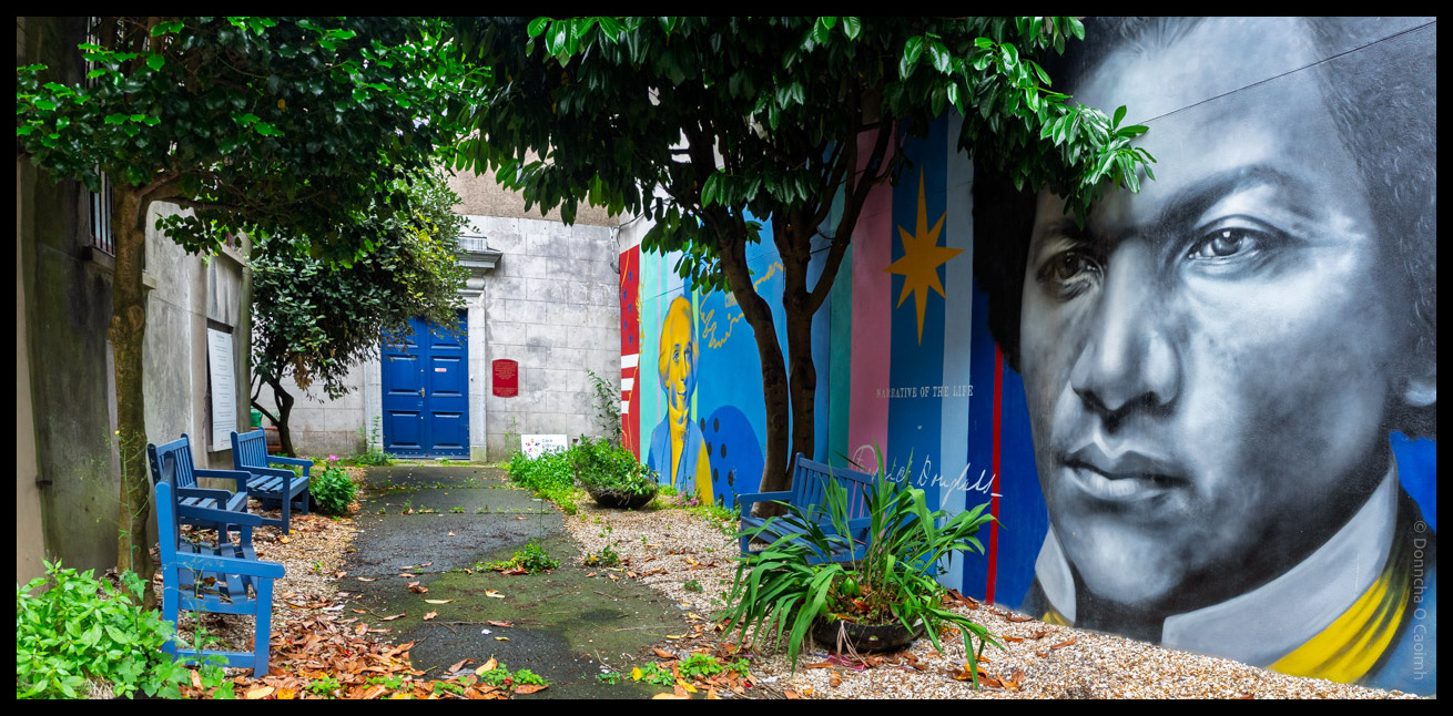 A vibrant avenue in Cork, Ireland, leads to the blue double doors of the historic Unitarian Church on Princes Street. The scene features lush green trees arching overhead, blue benches lining both sides of the path, and a colourful mural on the right wall depicting Frederick Douglass, with the words "Narrative of the Life" and a yellow star on a blue and pink striped background. The ground is scattered with leaves and gravel, and the atmosphere is peaceful and inviting.