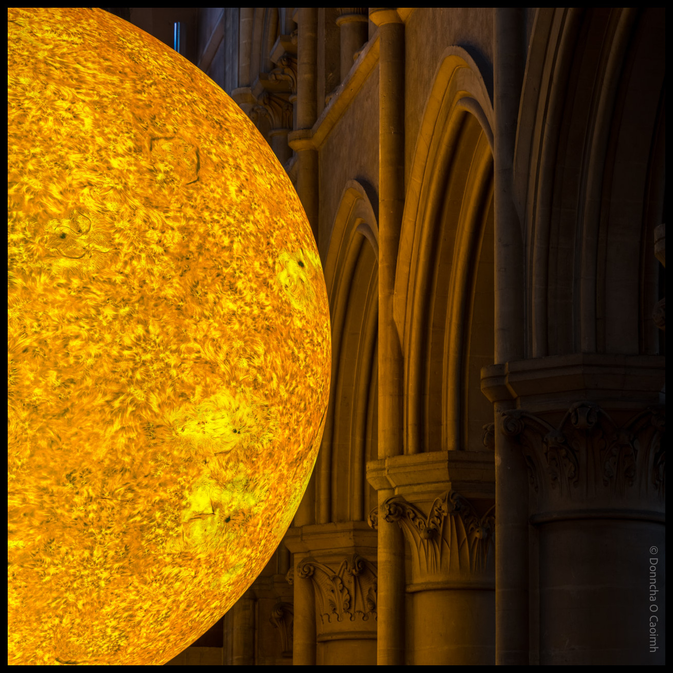 A striking photograph showing a large, illuminated model of the Sun with detailed surface textures, positioned inside a grand, arched stone cathedral; the image is warm-toned and atmospheric.