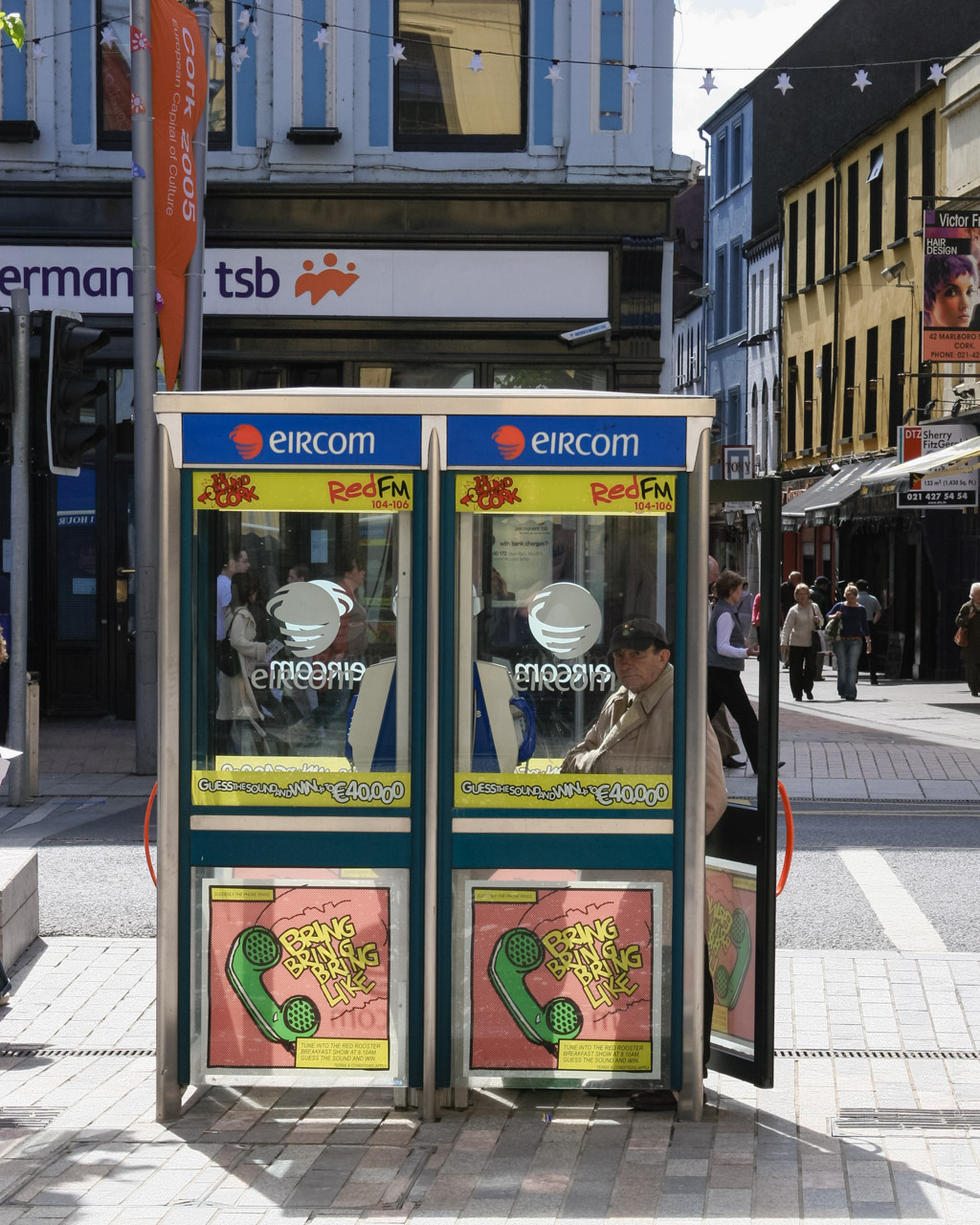 A pair of classic Eircom phone booths stand side by side on a busy pedestrian street in Cork, Ireland, with colourful "BRING BRING BRING LIKE" posters featuring green telephones on the lower panels, RedFM 104-106 and Eircom branding on the upper panels, a bank sign, "permanent tsb", in the background, and people walking by on a sunny day.

