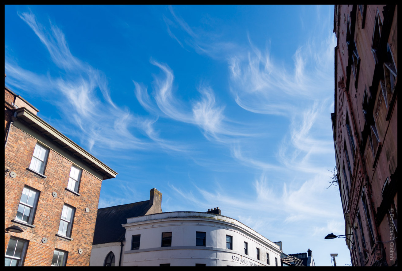 Urban street scene showing historic brick and white buildings in Cork City with wispy white clouds against a bright blue sky, featuring a distinctive curved white building with "CATHOLIC YOUNG" visible on its facade.
