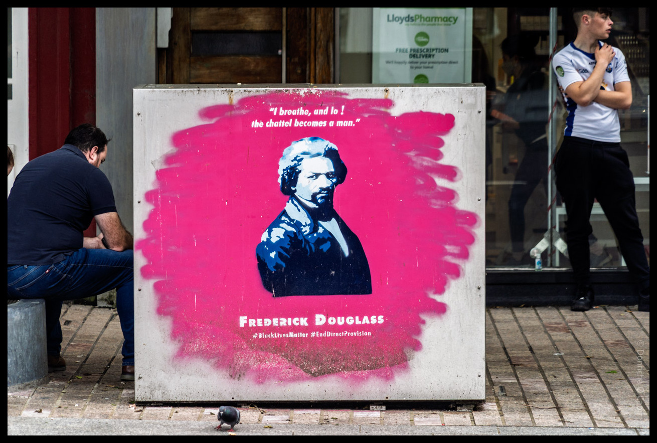 Street art mural on a city utility box featuring a painted portrait of Frederick Douglass on a vibrant pink background, with the quote "I breathe, and lo! the chattel becomes a man." 
Teh text below reads: "FREDERICK DOUGLASS #BlackLivesMatter #EndDirectProvision". Two people are visible nearby, one sitting and one standing, with a LloydsPharmacy storefront in the background and a pigeon on the pavement in front.
