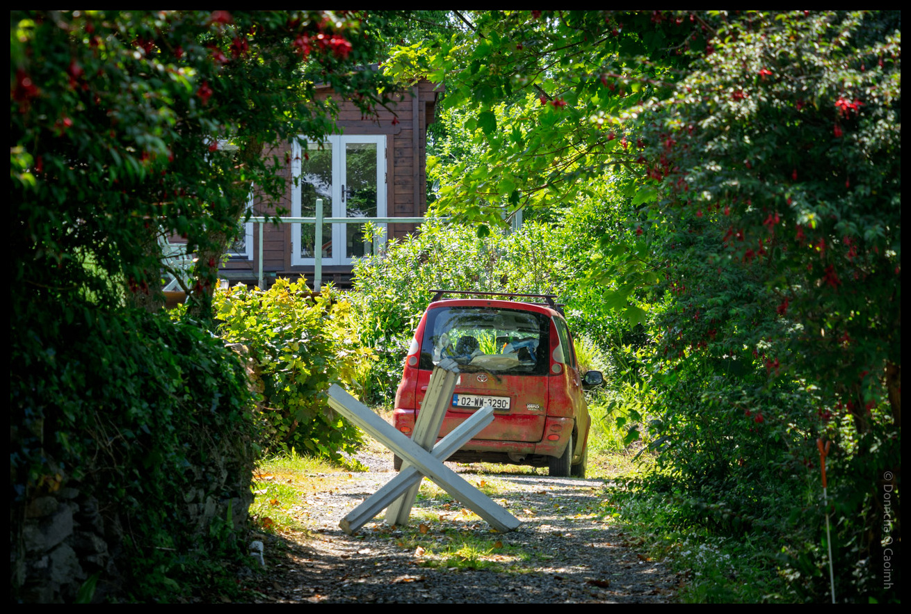 A red Toyota van parked on a gravel driveway beside a wooden house with white balcony railings, surrounded by lush green foliage and trees with some autumn colours, with wooden crossed barriers blocking the path.