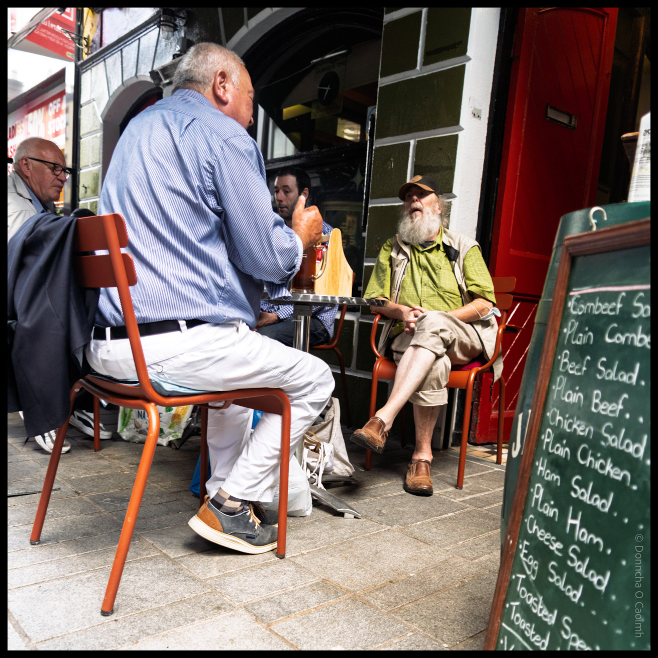 Street scene showing people seated at outdoor tables outside The Long Valley bar in Cork, with a green chalkboard menu displaying "Lombard St Plain Comb, Beef Salad, Plain Beef, Chicken Salad, Plain Chicken, Ham Salad, Plain Ham, Cheese Salad, Egg Salad, Toasted Special, Toasted"