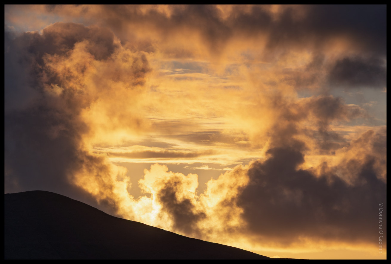 Dramatic sunset sky over the Dingle Peninsula in County Kerry showing golden and orange clouds illuminated against dark storm clouds with the silhouette of a hill in the foreground.
