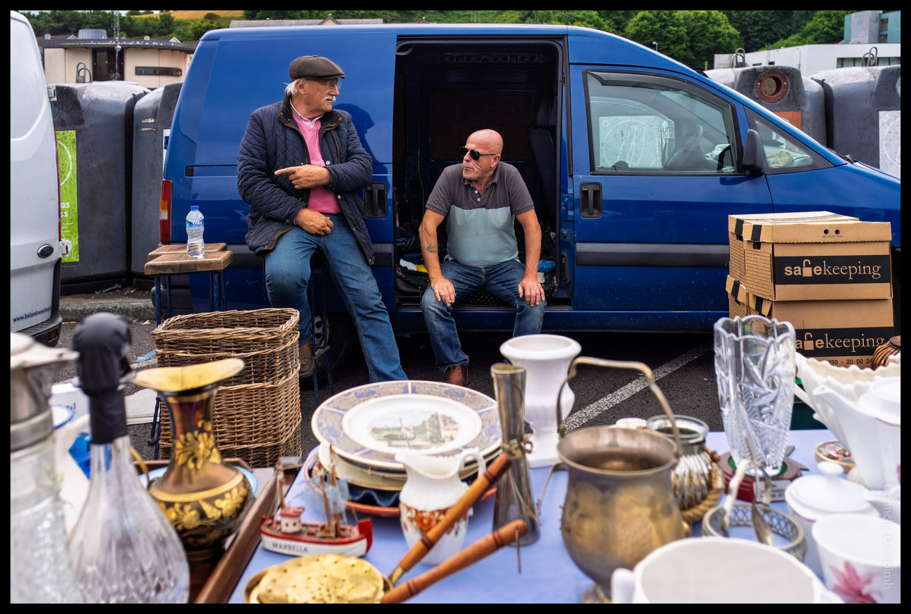 Two men sitting in the back of a blue van at a car boot sale or market in Bantry, County Cork, surrounded by various antiques and household items including brass vessels, wicker baskets, plates, and "safekeeping" boxes.