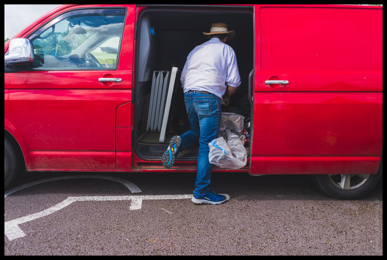 Man in brown hat, white shirt and jeans loading or unloading items from the sliding door of a bright red van at Bantry market, with plastic bags and materials visible inside the vehicle.
The van is parked on top of an accessible parking space.