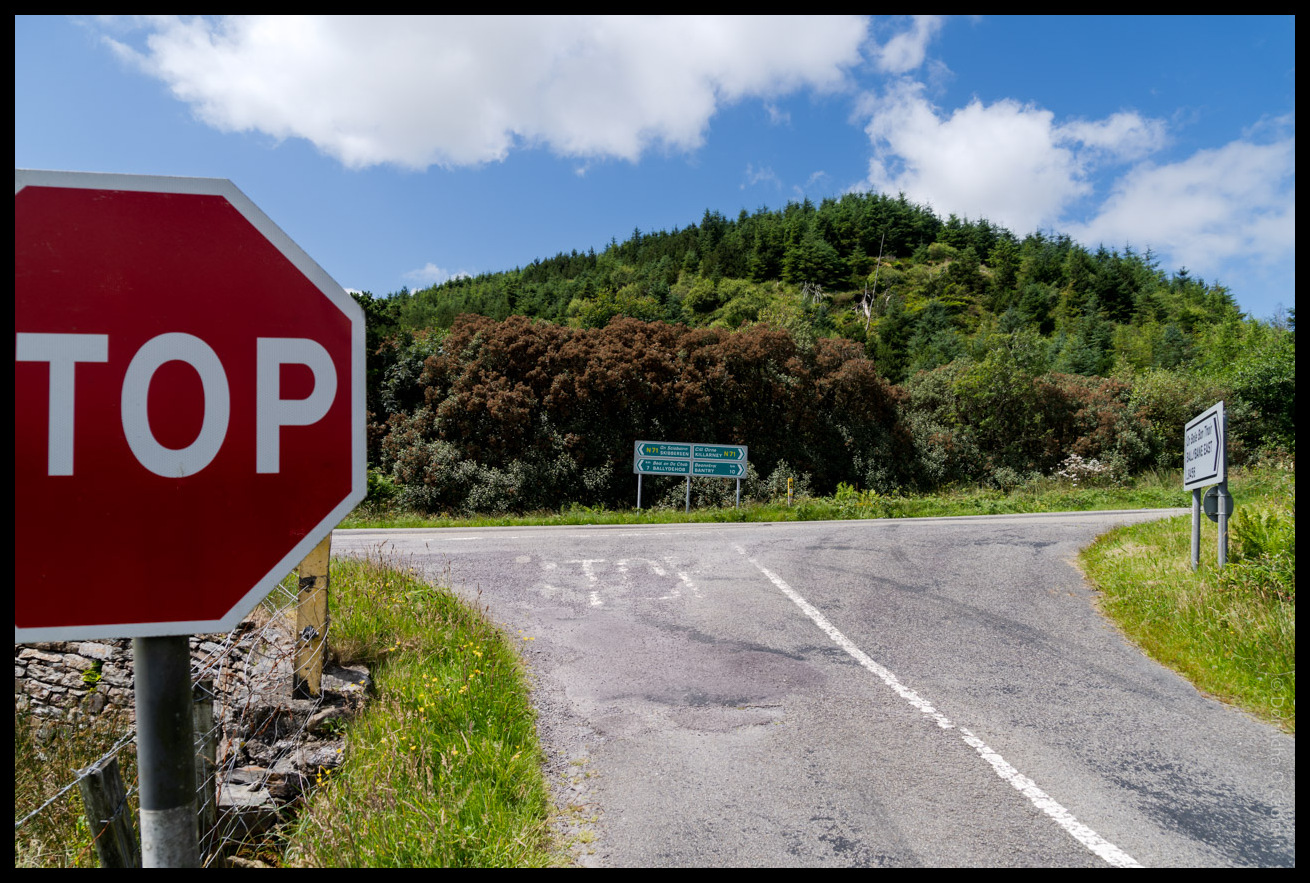 Rural road photograph: A red octagonal STOP sign prominently positioned on the left side of a narrow country road intersection, with green directional road signs visible in the background pointing to various destinations, surrounded by lush green hills covered in mixed forest and heather, stone walls, and verdant Irish countryside under a partly cloudy blue sky.
