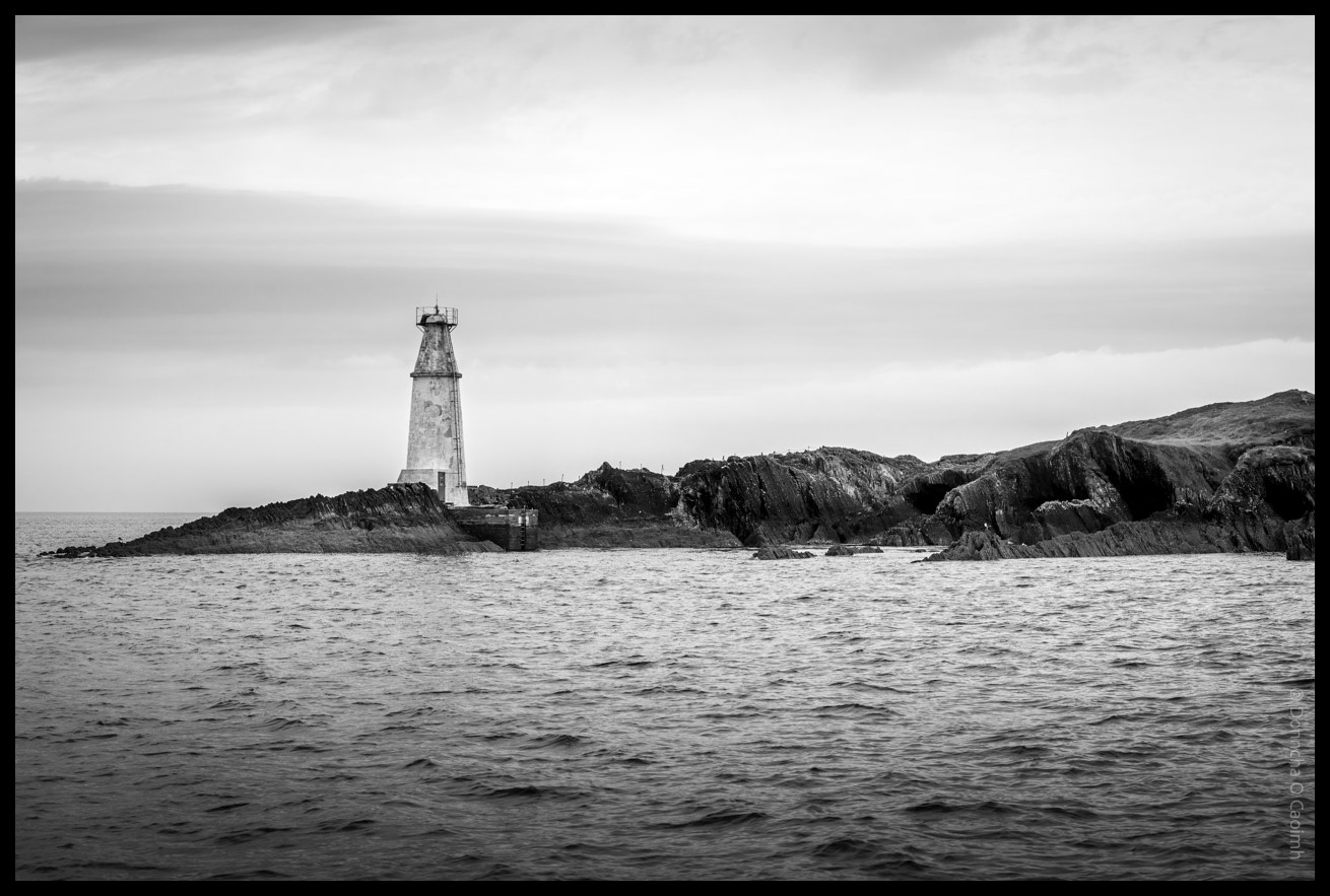 Black and white photograph of Copper Point Lighthouse standing on a rocky coastline with choppy waters in the foreground and overcast sky above.