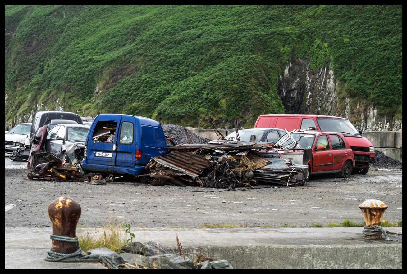 Scrapyard of abandoned and damaged cars including a blue van and red vehicles piled with corrugated metal and debris on Cape Clear Island, with lush green hills in the background and rusted bollards in the foreground.