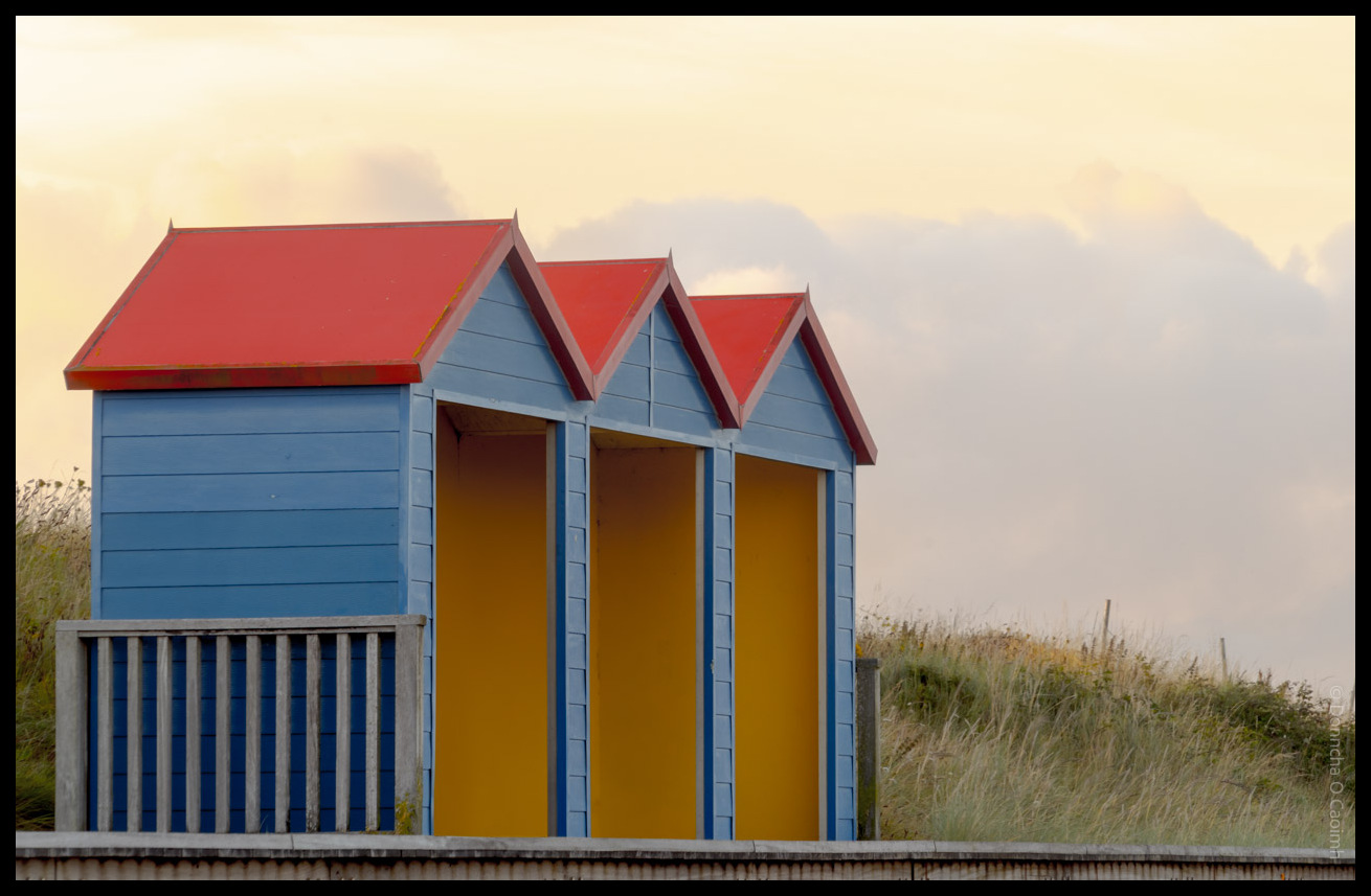 Colourful beach huts with red roofs, blue walls and bright yellow doors lined up behind a blue and white striped windbreak among coastal grasses at Youghal beach, County Cork, under a golden evening sky.