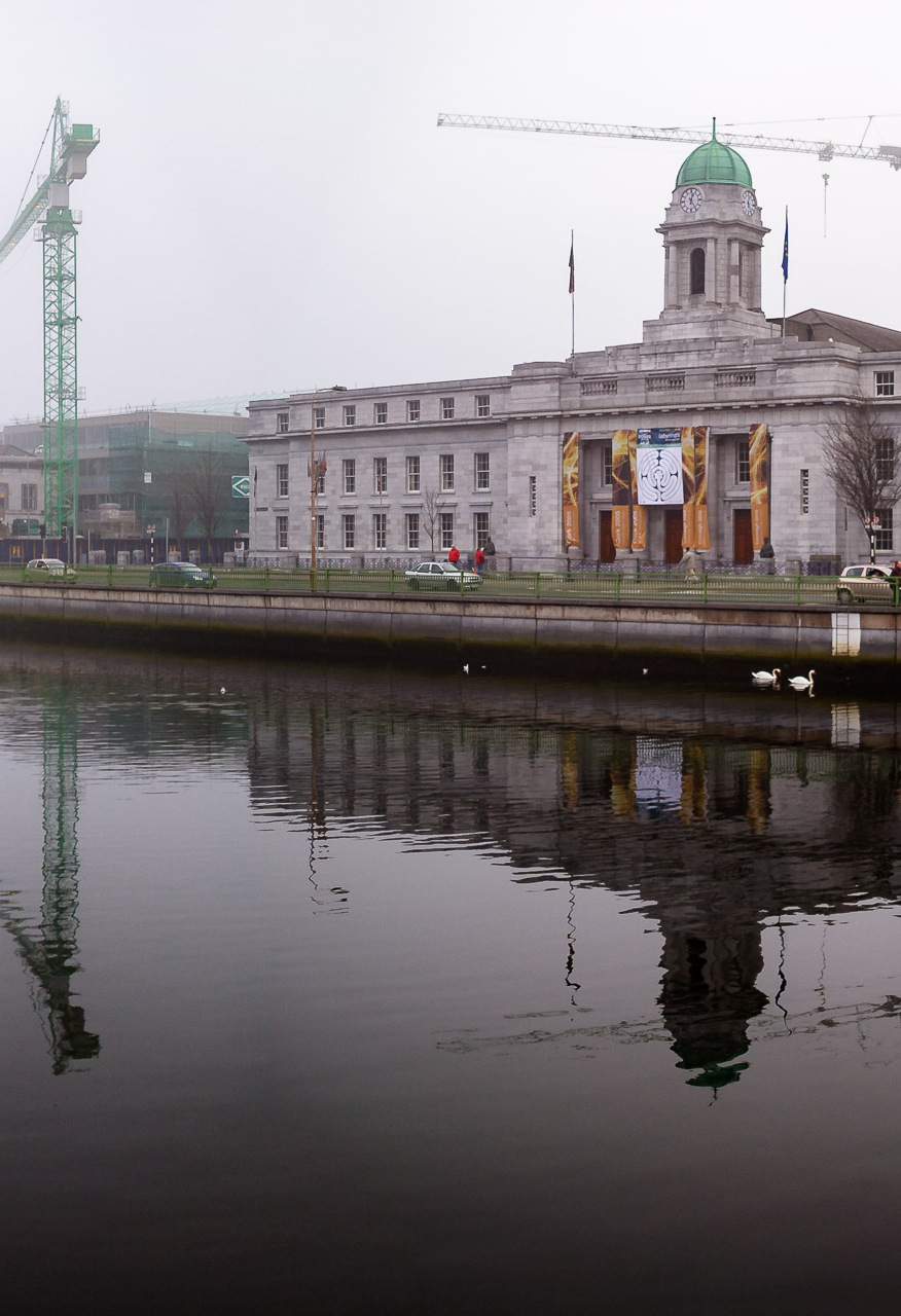 2005 photograph of Cork City Hall with its distinctive green dome reflected in the River Lee, showing construction cranes and the area before The Elysian development and modernisation, with swans visible on the calm water (2/3)