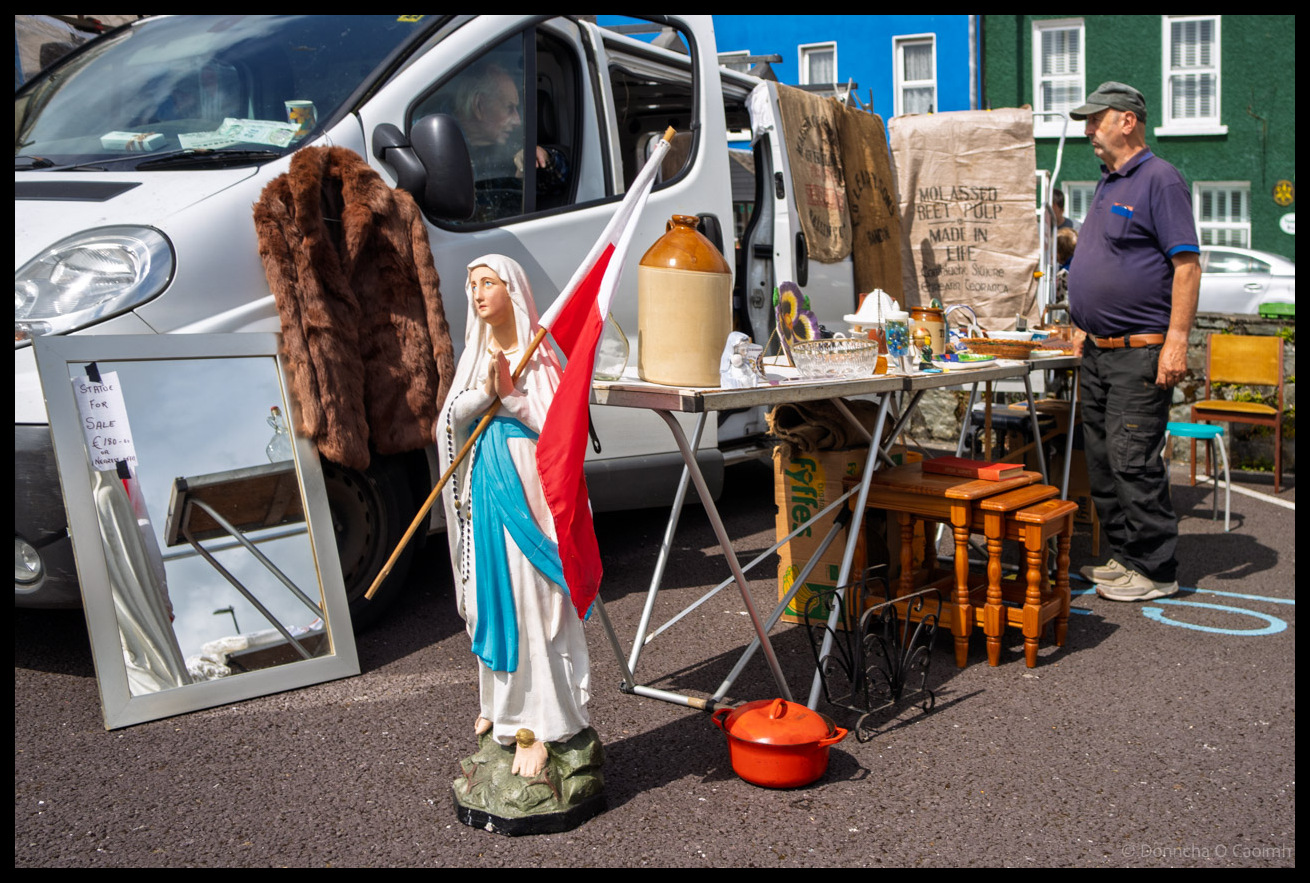 Market vendor at Bantry displaying an eclectic mix of items including a religious statue holding a red and white Cork flag, fur coat, ceramic jug, wooden furniture and various antiques spread around his van, County Cork.
