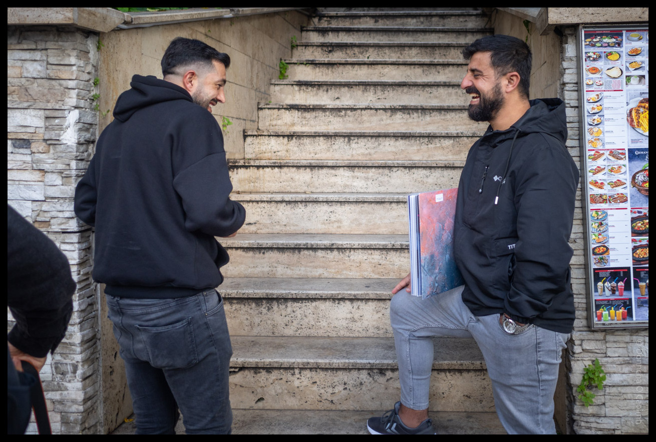 Two men in black hoodies and jeans having a friendly conversation while sitting and standing on concrete steps next to a restaurant menu display in Istanbul, Turkey.