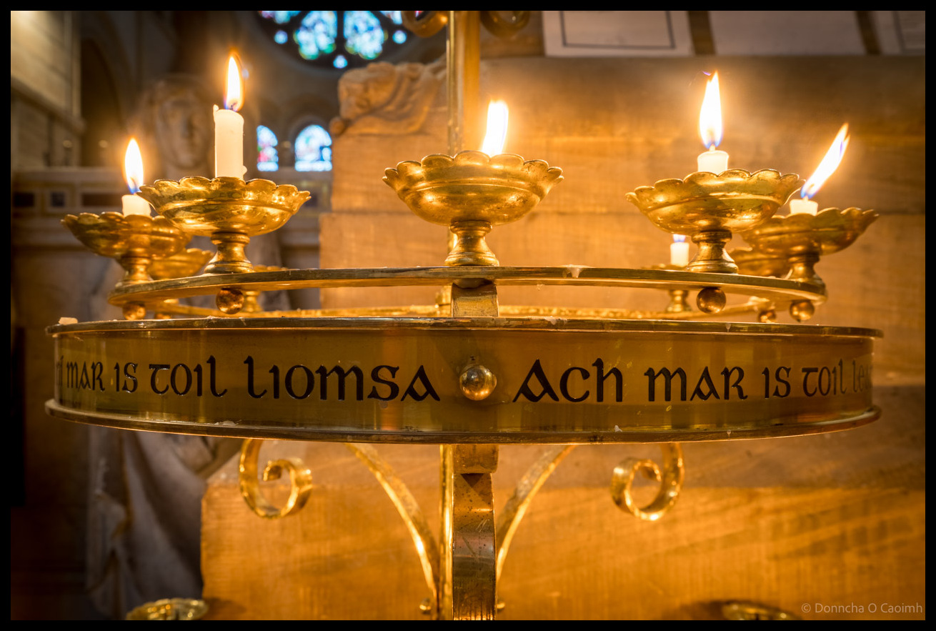 Ornate brass candle stand with lit white candles in decorative holders, inscribed with Irish text "NI MAR IS TOIL LIOMSA ACH MAR IS TOIL LEATSA" inside St Fin Barre's Cathedral, Cork, with stained glass windows visible in the background