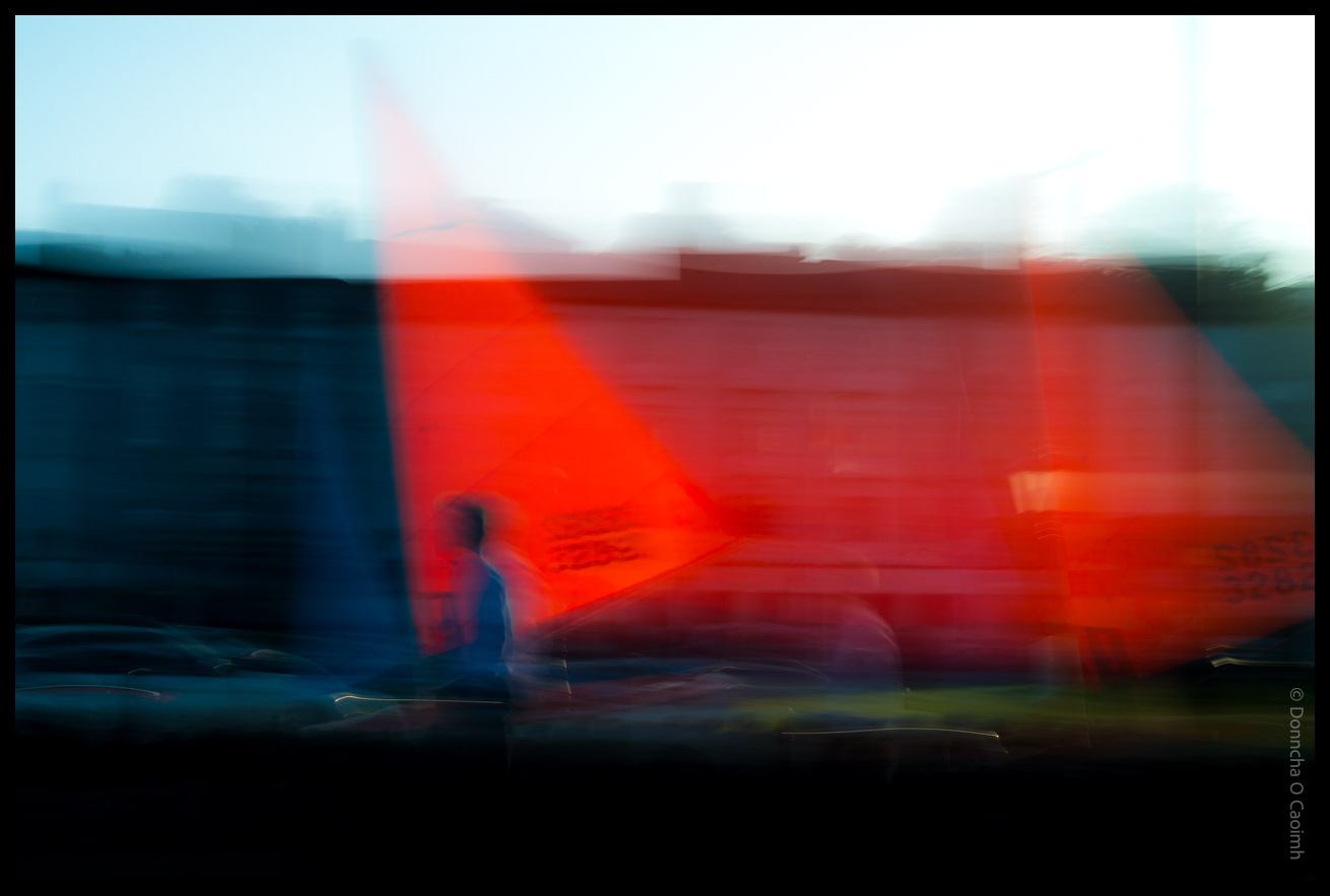 Abstract long exposure photograph showing motion blur of a person walking in front of a bright red boat sail in Kinsale harbour, County Cork, creating streaks of red, blue and white colours.