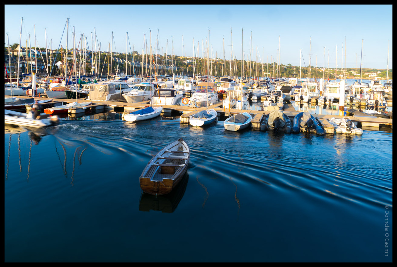 Bustling marina in Kinsale showing numerous sailing boats and yachts moored at wooden pontoons with a small rowing boat in the foreground on calm blue water, backed by tree-covered hills under a clear sky in County Cork