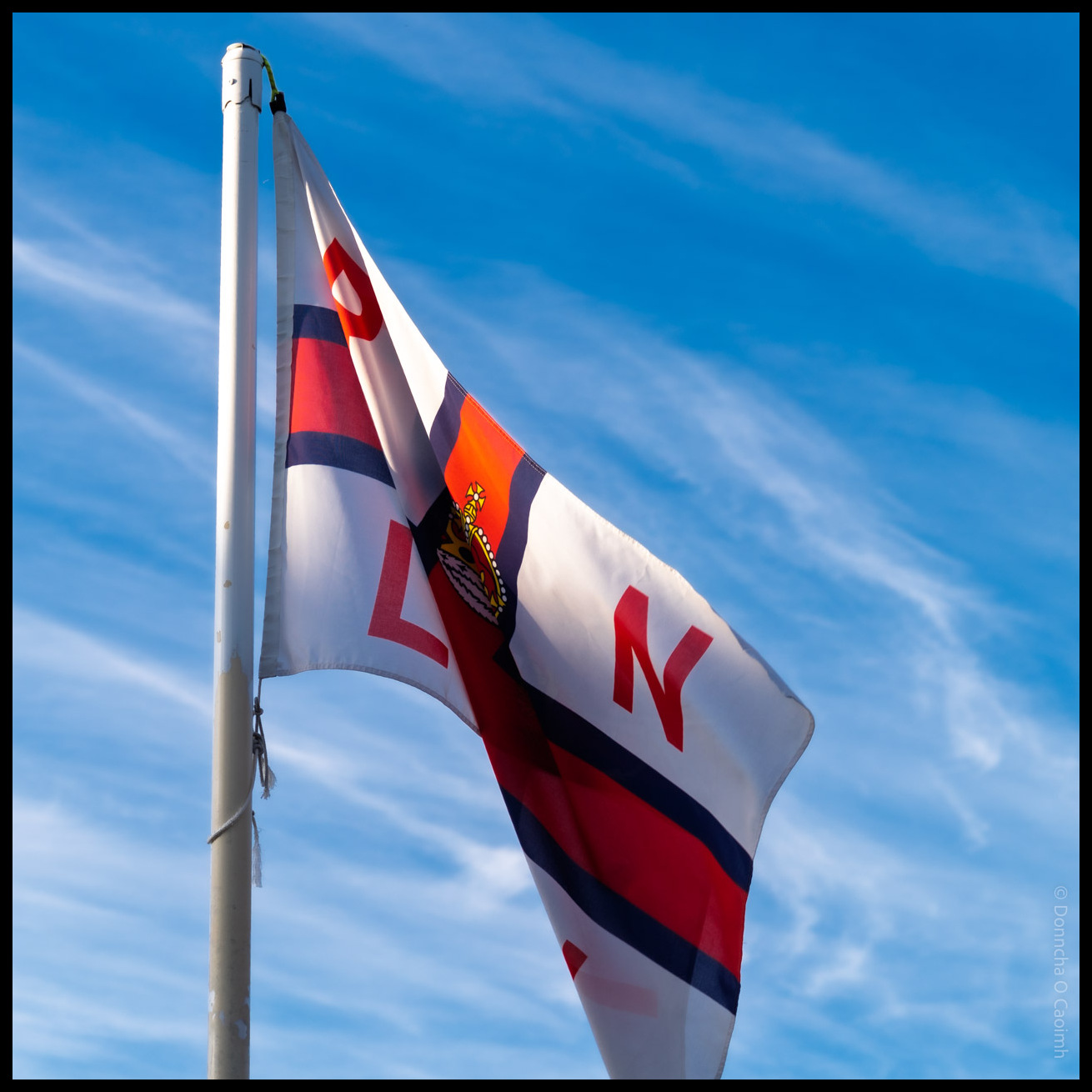 RNLI flag flying on a white flagpole against a bright blue sky with wispy white clouds in Ballycotton, County Cork.
