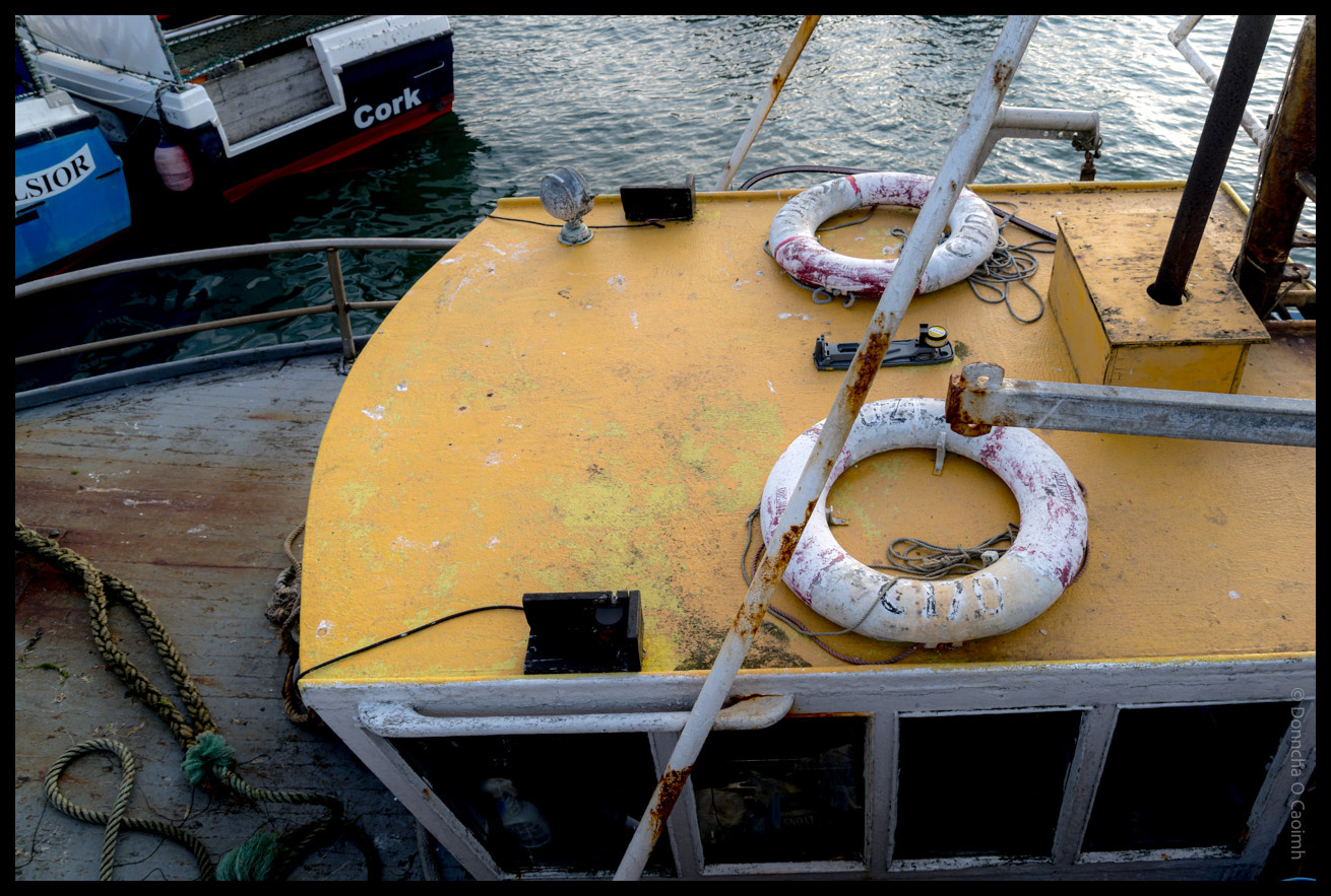 Close-up view of a yellow fishing boat deck showing life rings, rusty chains, and weathered equipment with other boats visible in Ballycotton harbour, County Cork