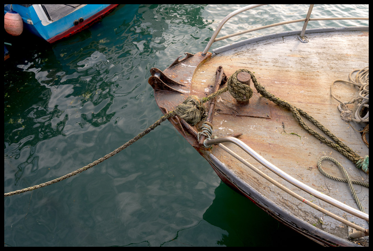 Fishing boat bow with heavy anchor chain, mooring ropes, and weathered wooden deck reflected in calm green harbour water in Ballycotton, County Cork