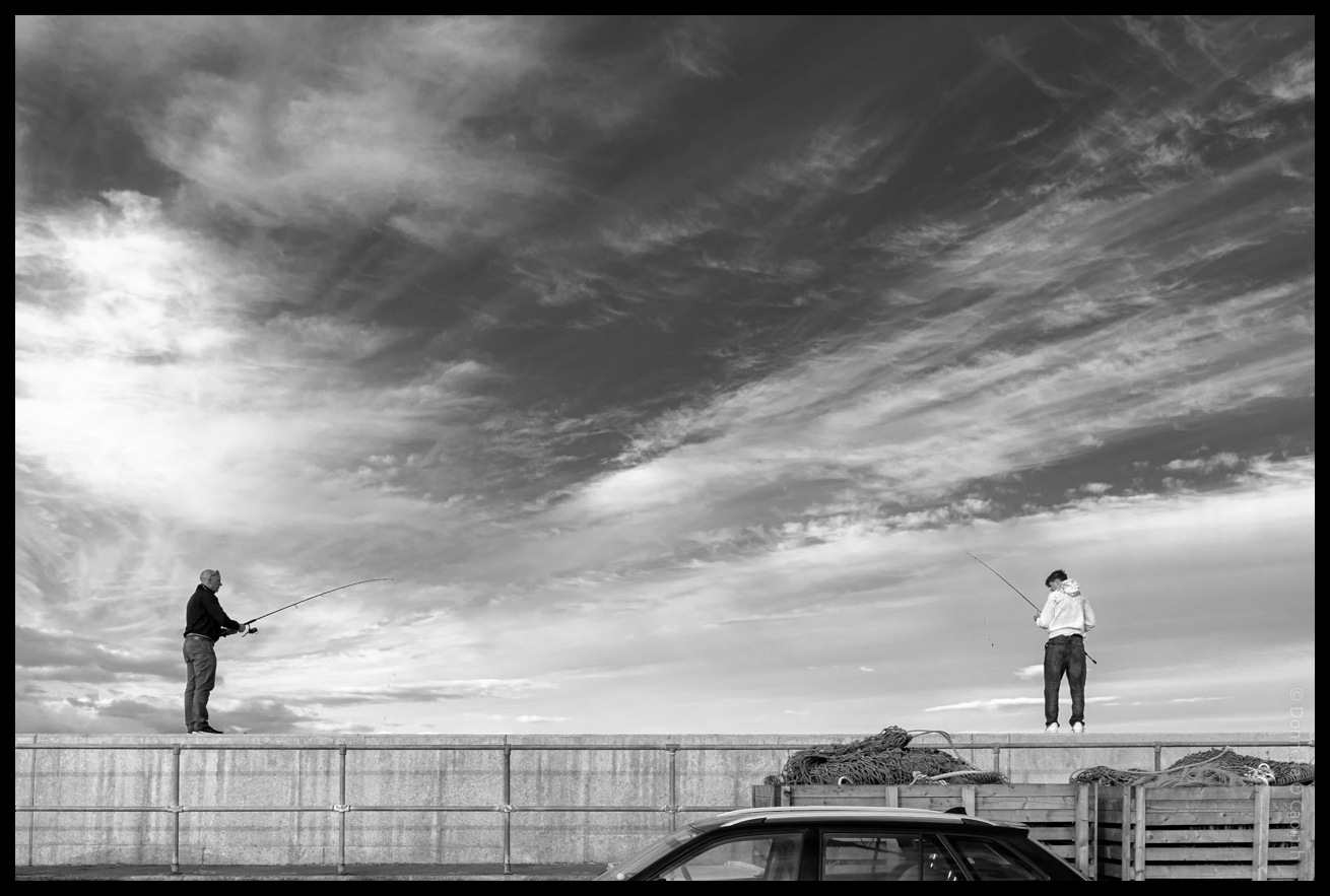 Black and white photograph of two people fishing from a concrete pier with dramatic cloudy sky overhead, fishing nets stacked on the pier and a car visible below in Ballycotton, County Cork.