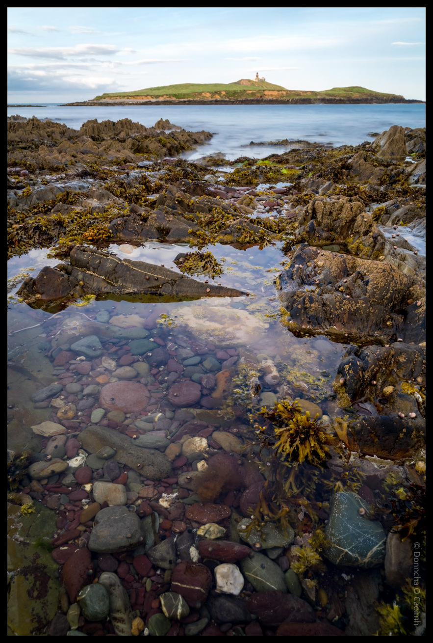 Rocky shoreline and tidal pools with colourful pebbles, seaweed and clear water in the foreground, with Ballycotton Lighthouse visible on its island in the background under a cloudy sky, County Cork.