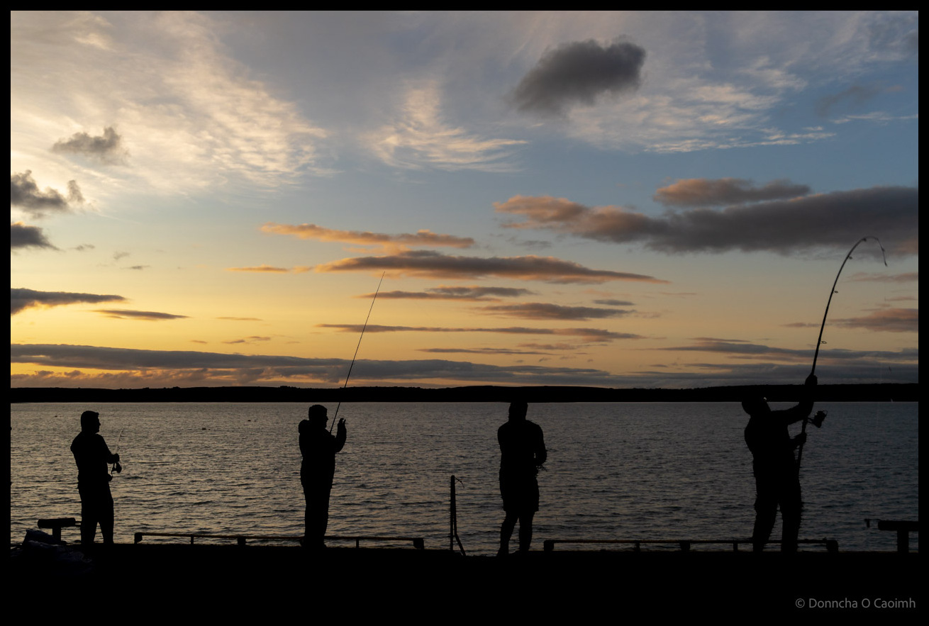 Silhouettes of four anglers fishing from a pier at sunset with dramatic cloudy sky and calm water in Ballycotton, County Cork