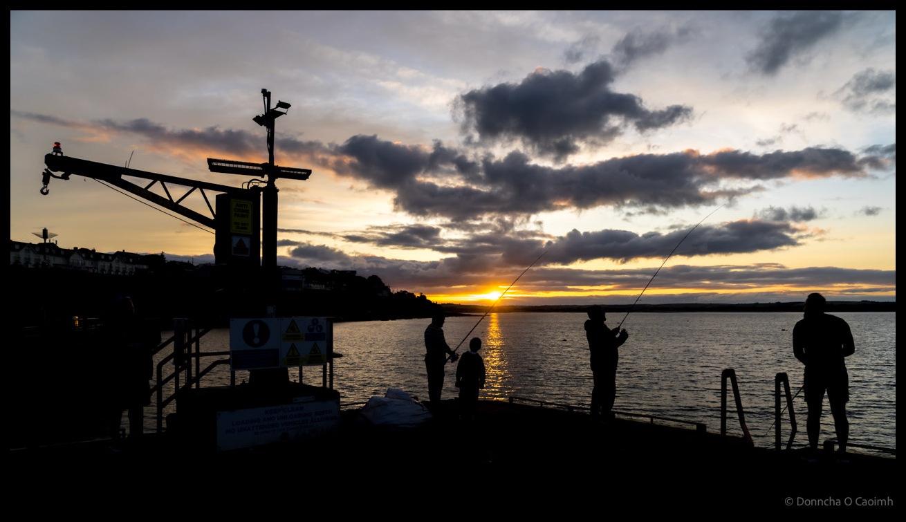 Sunset fishing at Ballycotton pier with harbour crane silhouetted against dramatic sky, anglers casting lines as sun sets over the water, County Cork